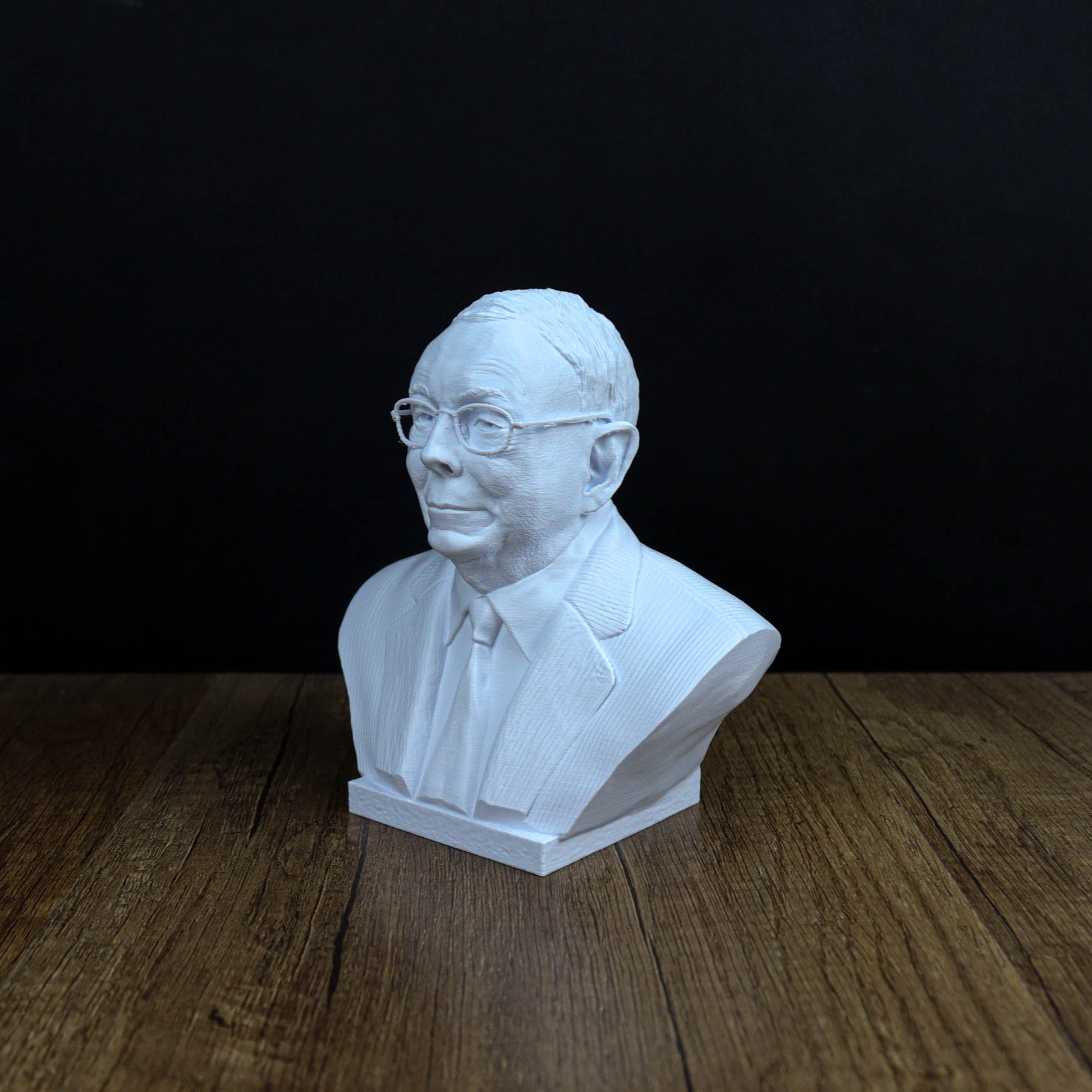 A white bust of a man wearing glasses and a suit is displayed on a wooden surface against a black background.
