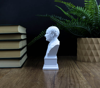 A white bust of a man is displayed on a wooden surface, surrounded by a stack of books and a potted plant.