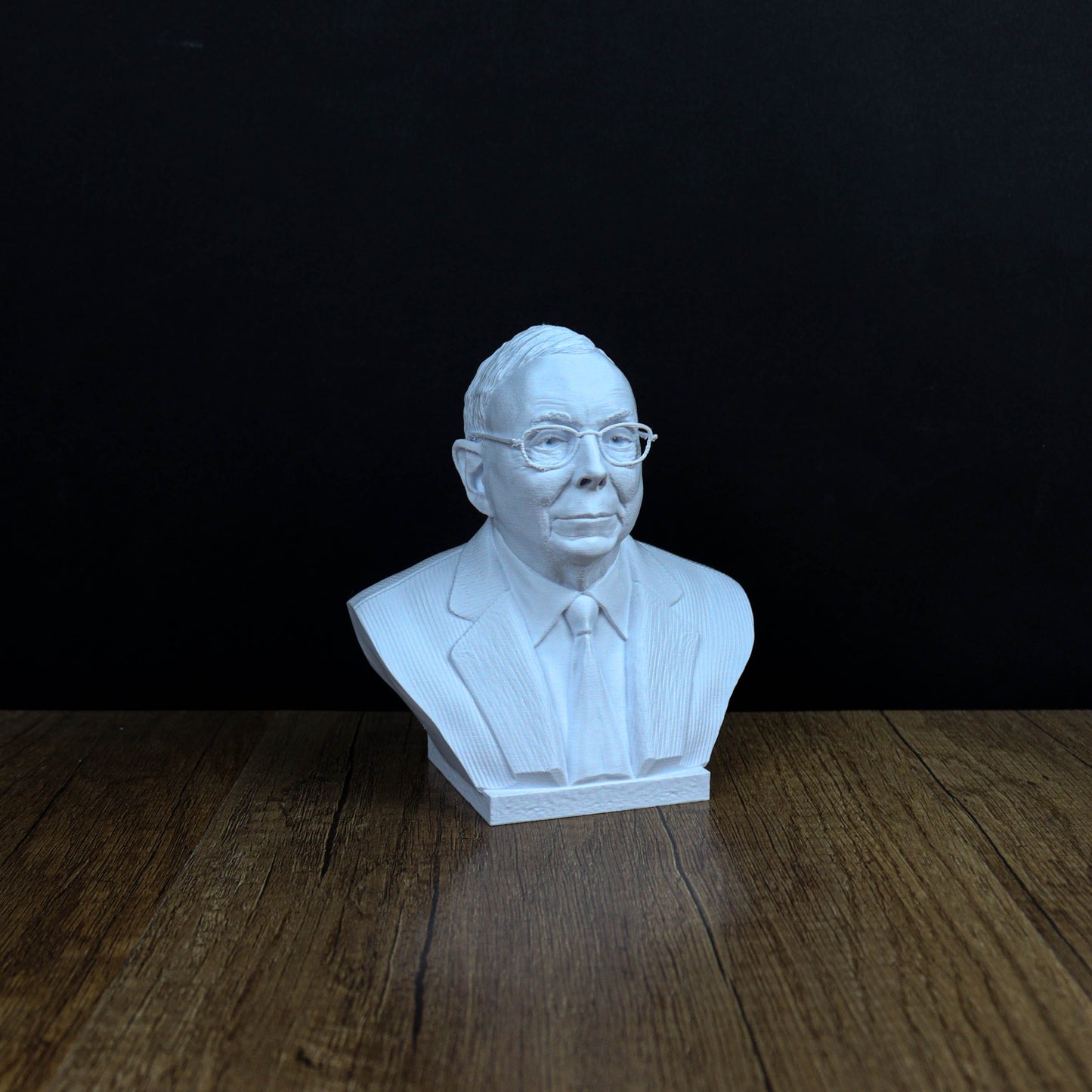 A white bust of a man wearing glasses and a suit is displayed on a wooden surface against a black background.