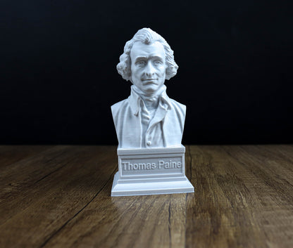 A white bust of a man, likely Thomas Paine, is displayed on a wooden surface against a dark background.