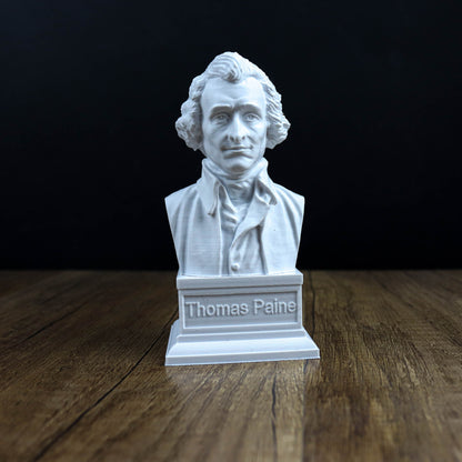 A white bust of Thomas Paine, a famous American author, is displayed on a wooden surface against a black background.