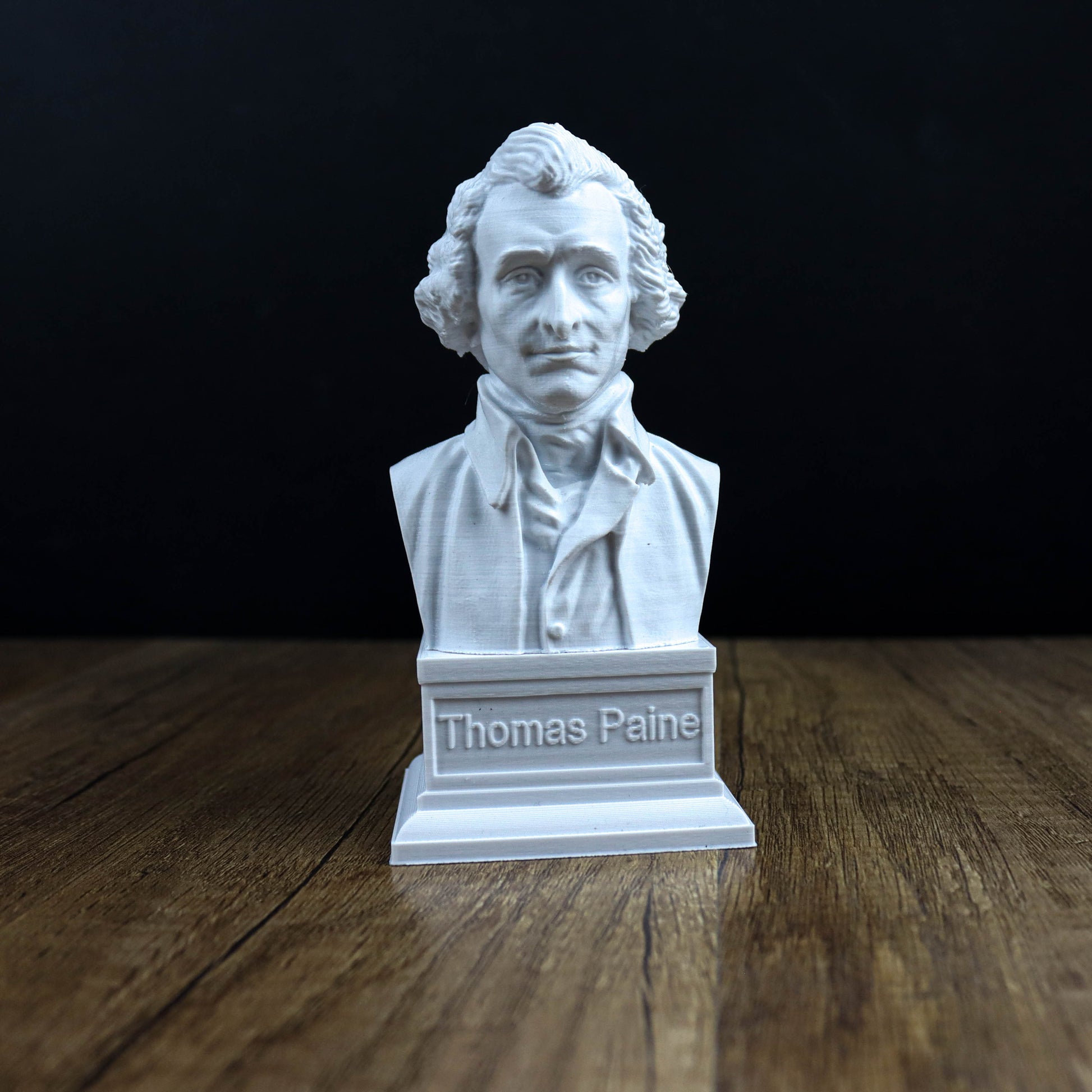 A white bust of Thomas Paine, a famous American author, is displayed on a wooden surface against a black background.